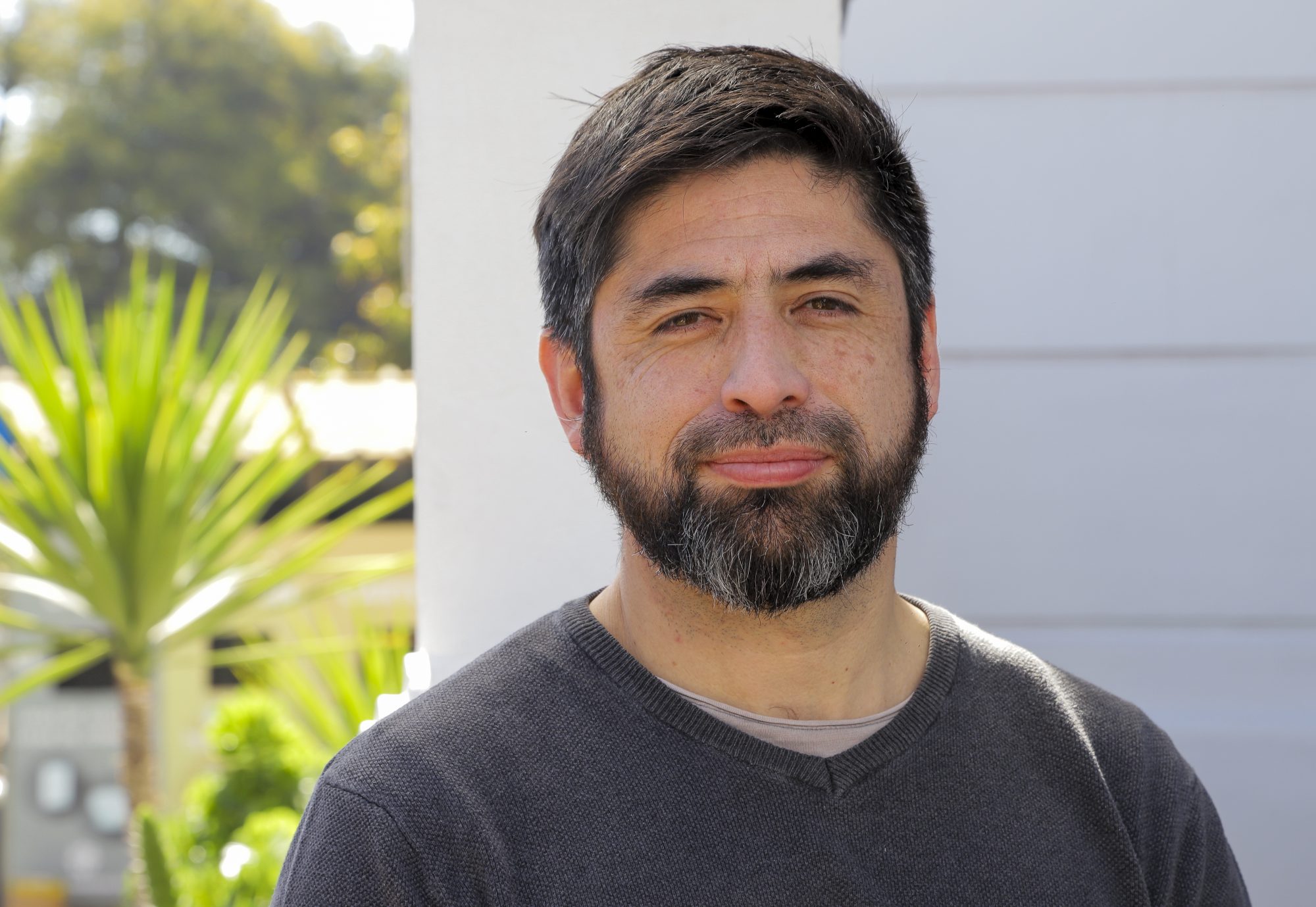 Man with short dark hair and gray beard standing outdoors near a white wall, with greenery in the background, looking toward the camera with a neutral expression.