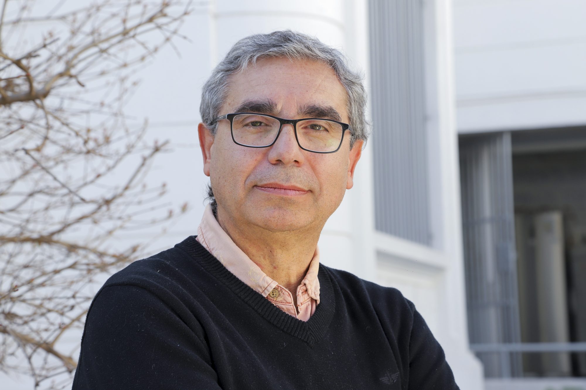 Portrait of a middle-aged man with gray hair and glasses standing outdoors in front of a light building.