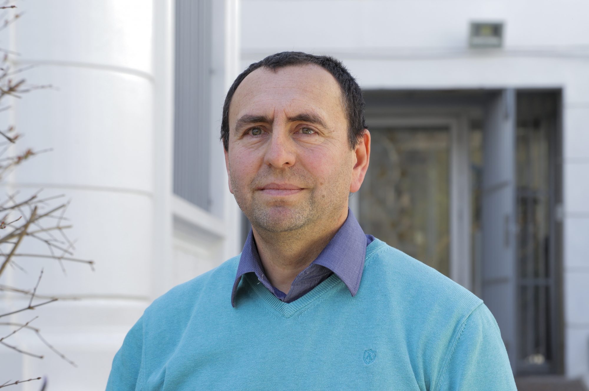 Portrait of a man outdoors wearing a turquoise sweater and blue collared shirt, standing in front of a white building.