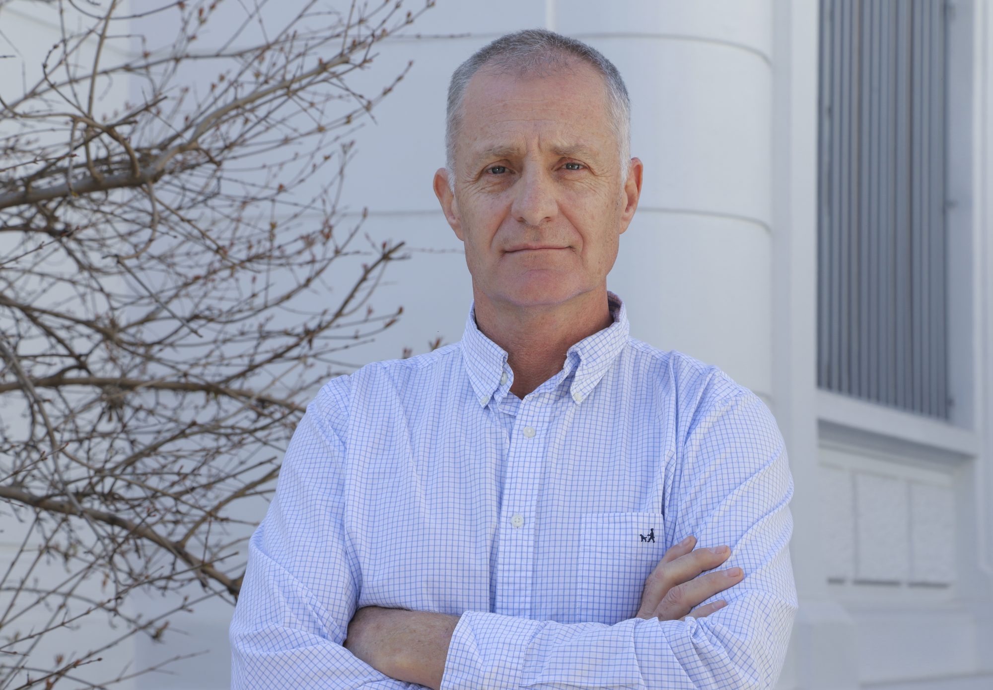 Portrait of a middle-aged man with short gray hair standing outdoors with arms crossed, near a white building and leafless branches.