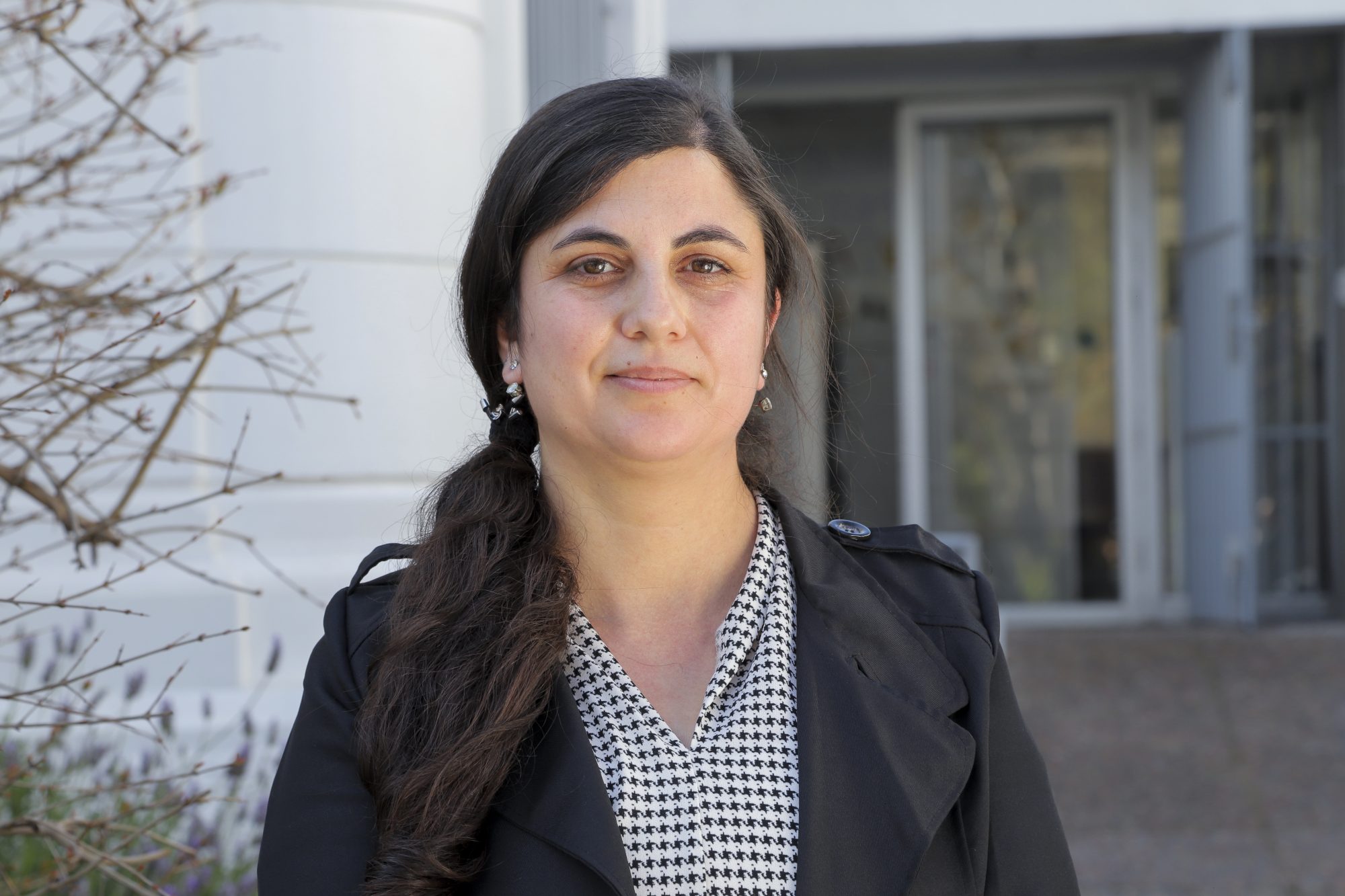 Portrait of a woman with dark hair in a black blazer and houndstooth scarf, standing outdoors in front of a building.