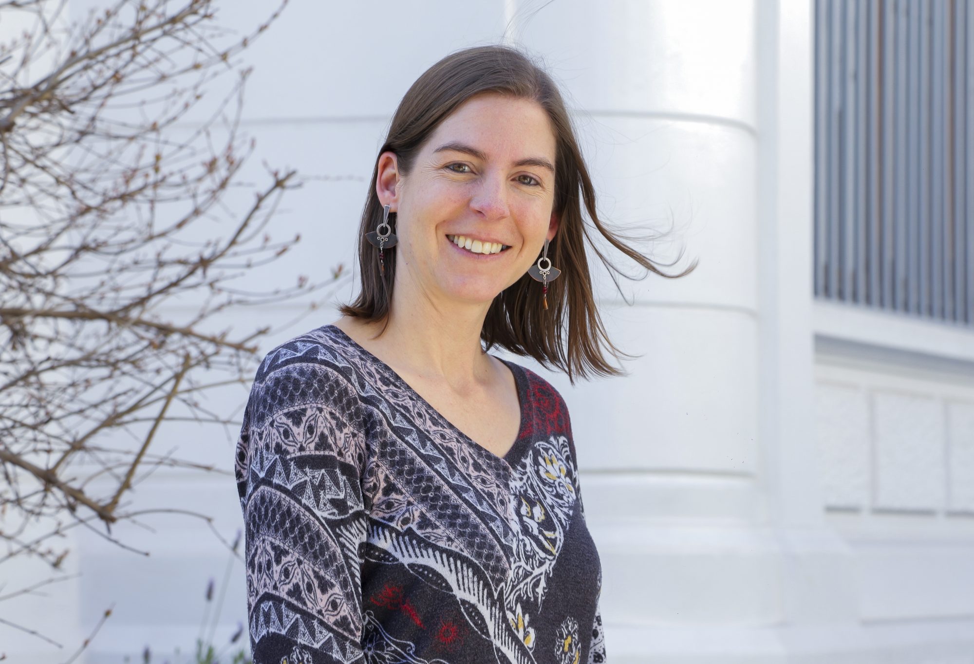 Smiling woman with shoulder-length brown hair in a patterned sweater, standing outdoors with bare branches on one side and a white building in the background.