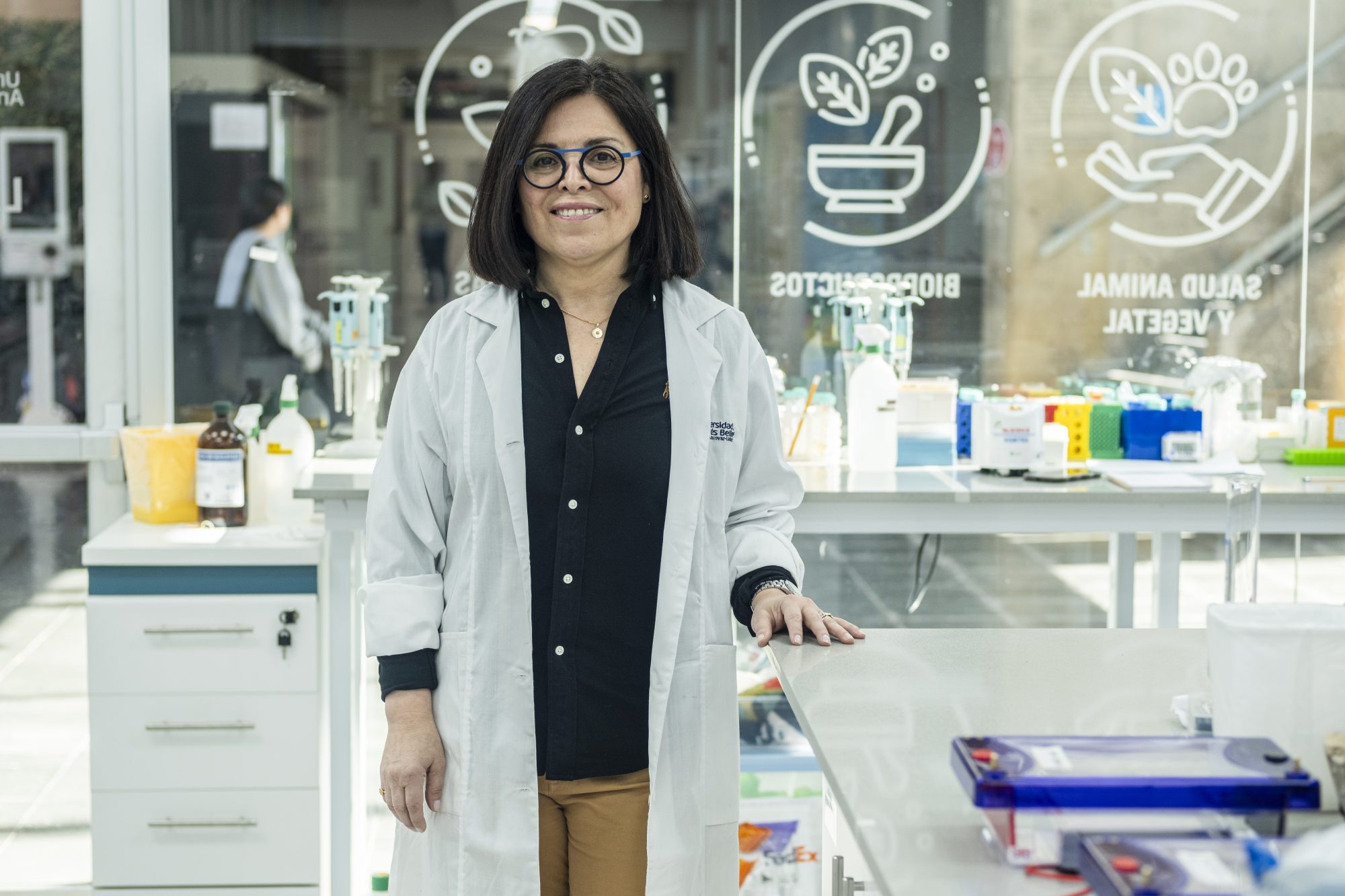 Female scientist in a white lab coat standing at a lab bench in a bright laboratory with equipment in the background behind glass panels.