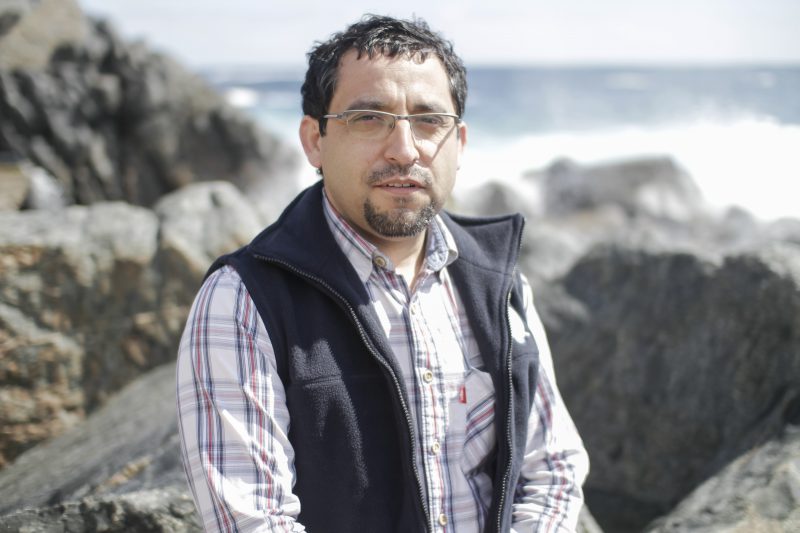Man wearing glasses and a navy vest over a plaid shirt sits among rocky coastal scenery with the ocean in the background.
