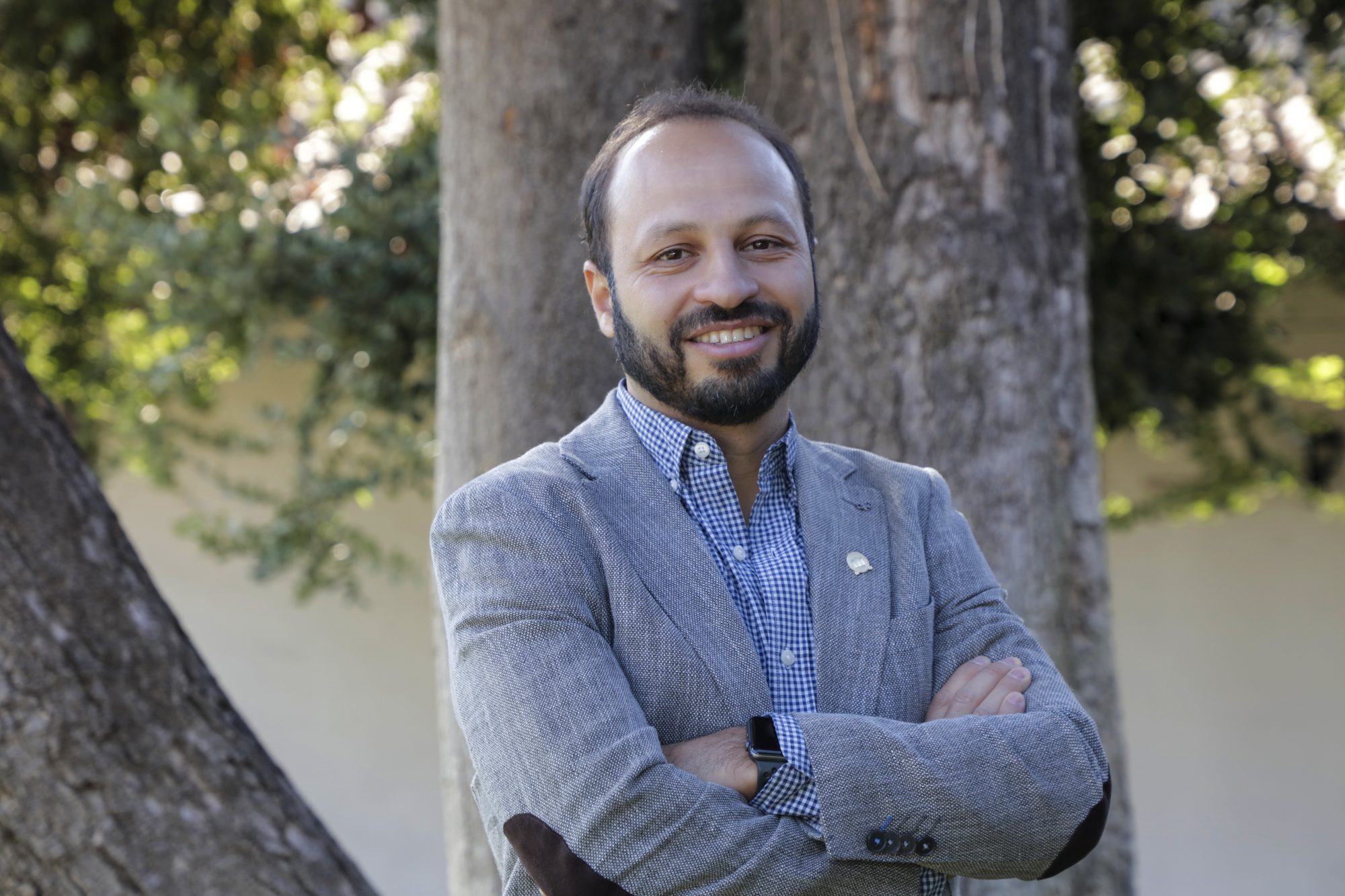 Smiling man in a gray blazer and blue checkered shirt stands outdoors with arms crossed in front of a tree trunk.