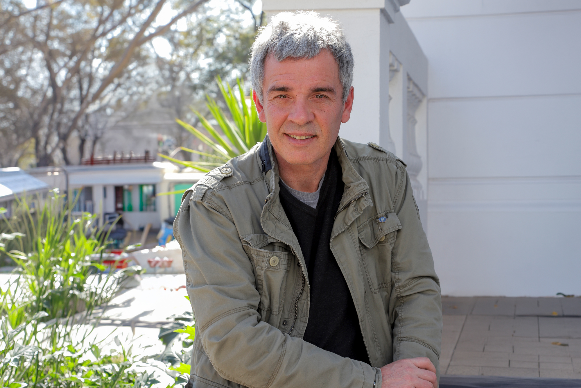 Casual portrait of a middle-aged man with gray hair, wearing a beige jacket, standing outdoors near a white building with plants in the foreground.