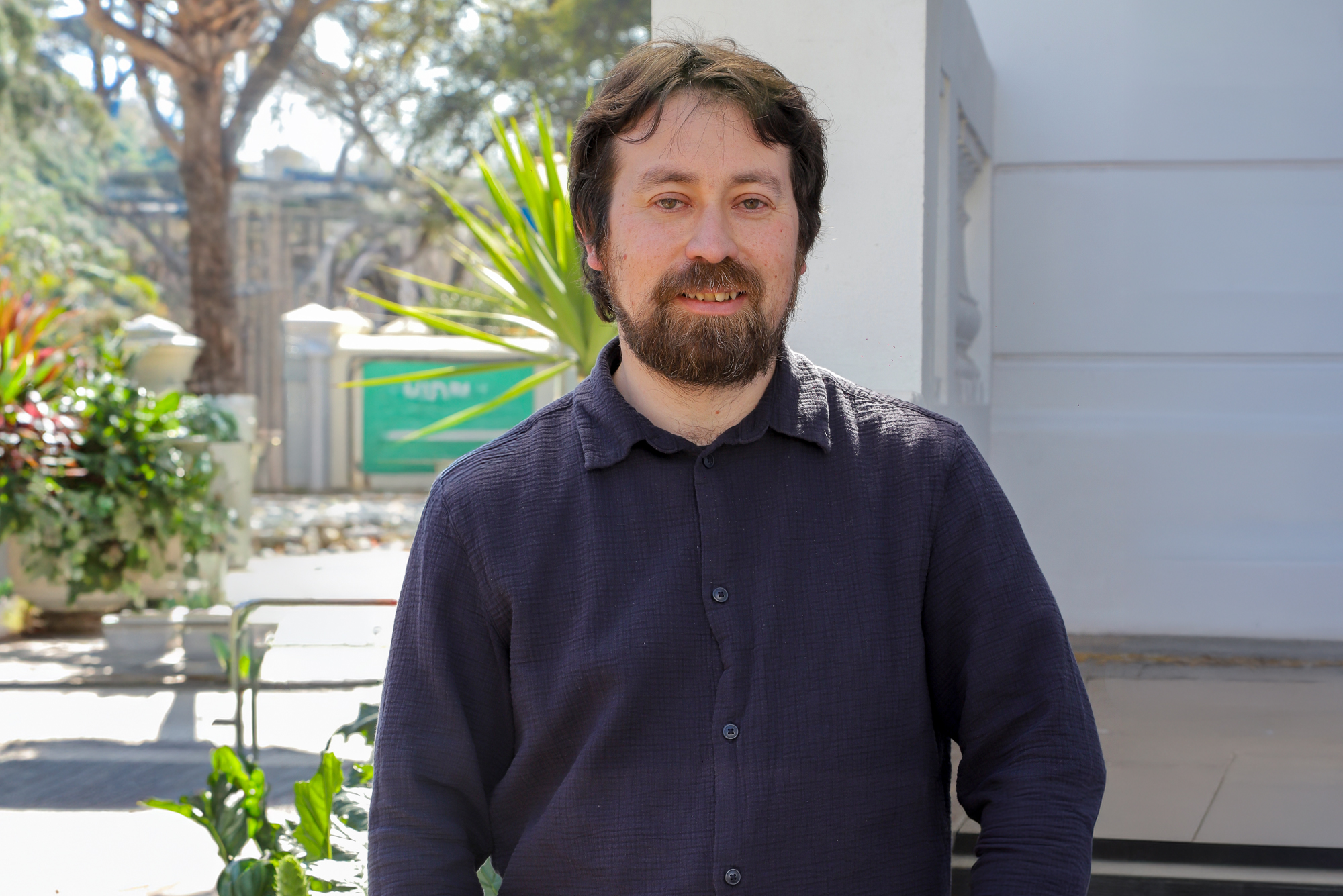 Portrait of a bearded man in a dark button-up shirt standing outdoors with greenery and a light wall behind him.