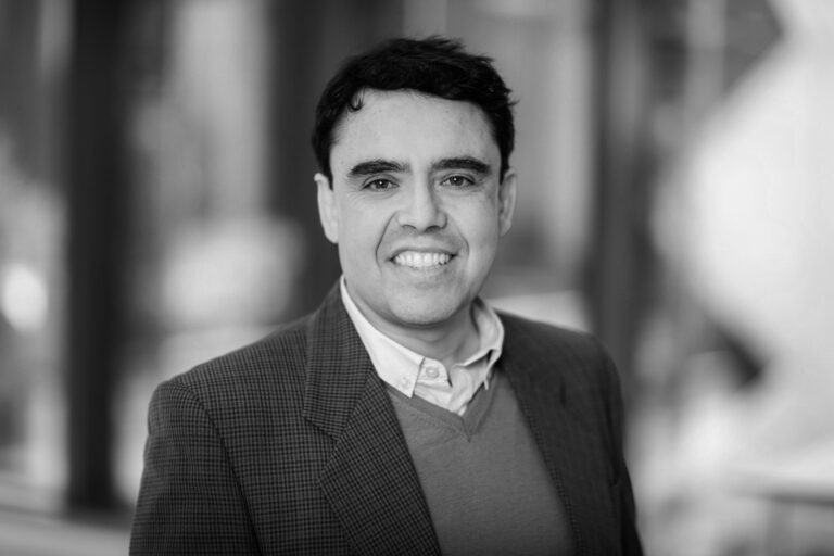 Professional head-and-shoulders portrait of a smiling man in a blazer and sweater, outdoor urban background (black and white).