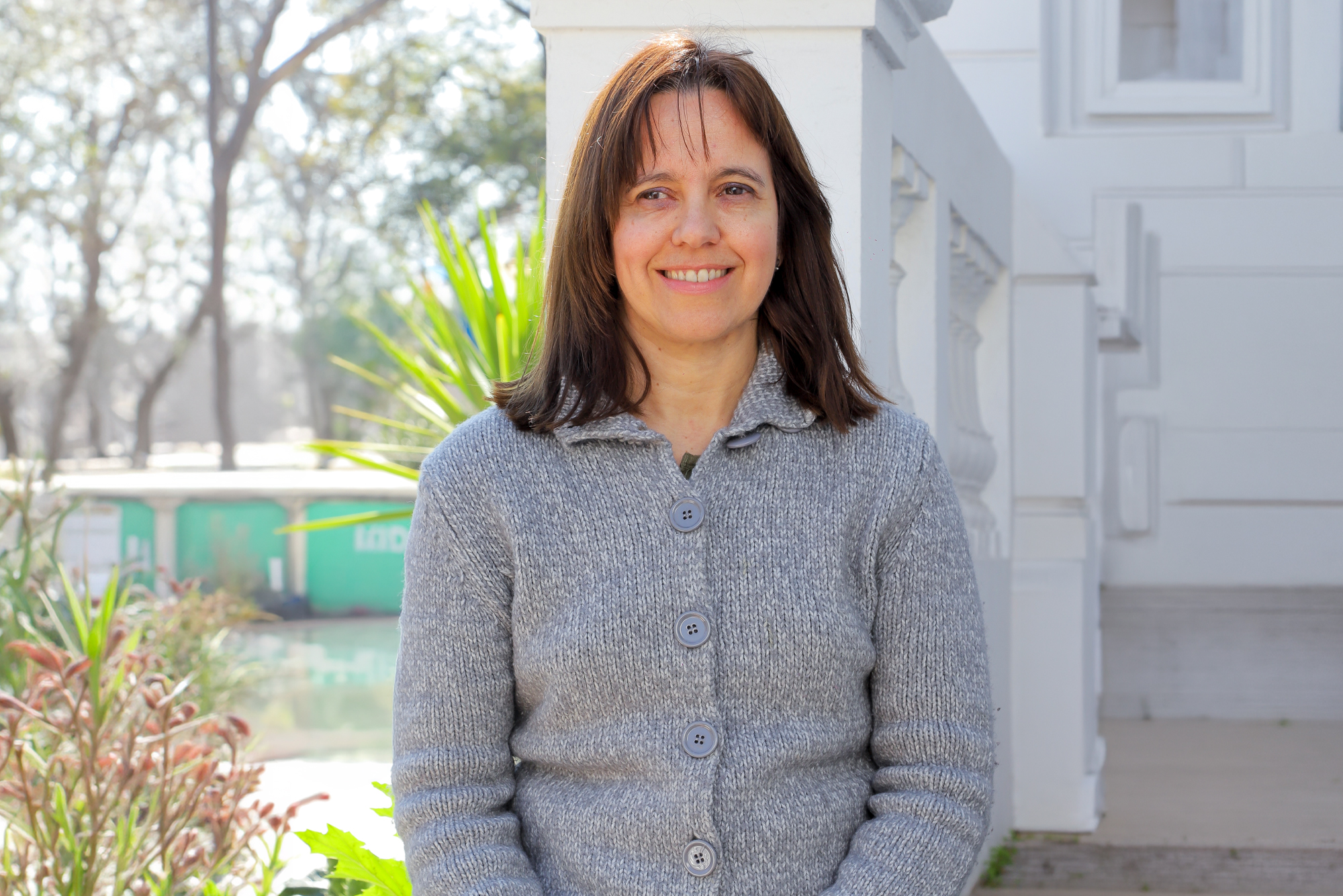 Smiling woman in a gray knit sweater standing outdoors beside a light-colored building with columns.