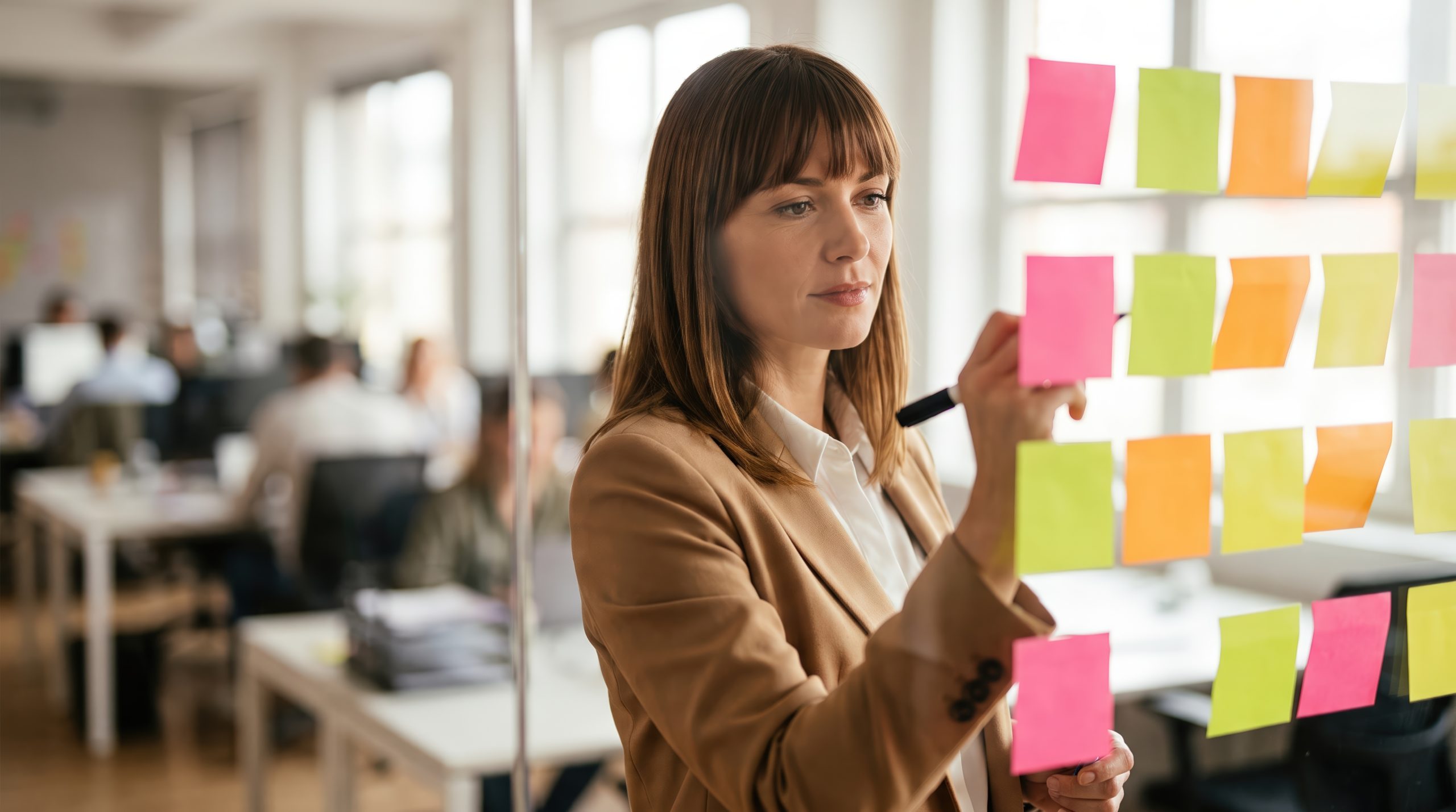 Female project manager writing on sticky notes on glass wall in modern office for agile workflow and strategy brainstorming