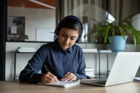 Una joven anotando en un cuaderno frente a una computadora en un entorno de estudio, concentrada en su trabajo.