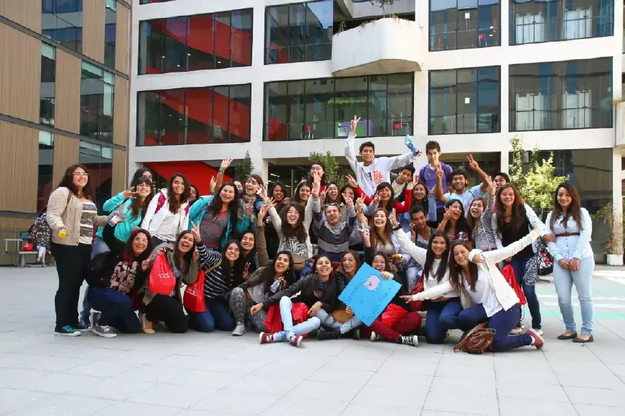 Un grupo de jóvenes posando animadamente para una fotografía en el patio de una universidad, todos sonrientes y mostrando señales de camaradería y alegría.